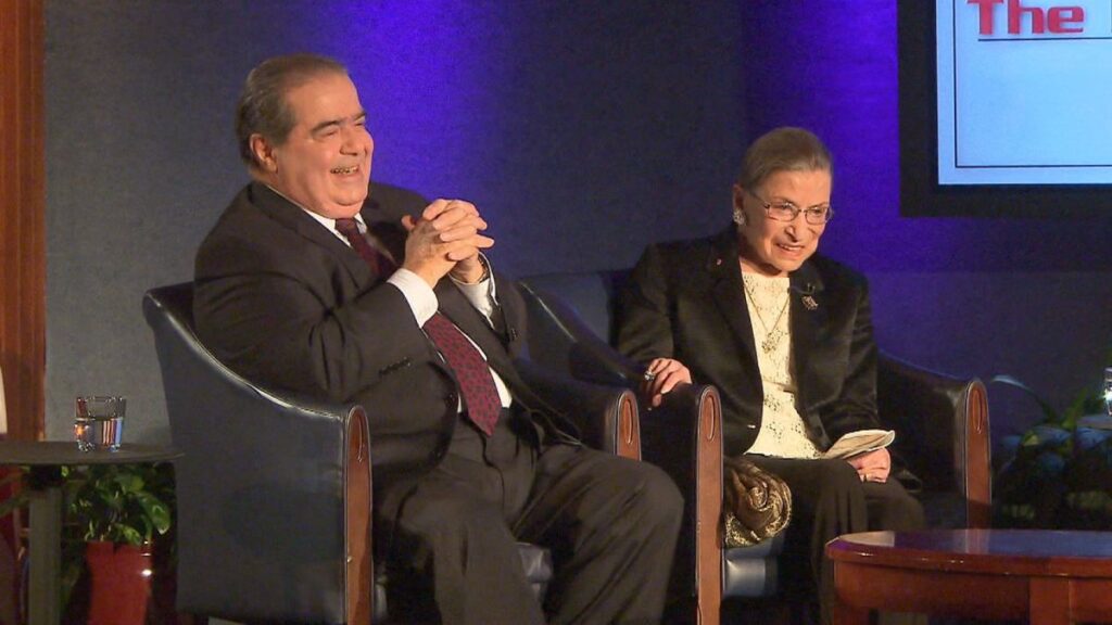 Supreme Court Justices Antonin Scalia and Ruth Bader Ginsberg seated next to each other during an interview. Both are smiling at an unseen person.